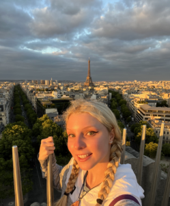 Mary is standing on a building with a view of Paris, France behind her. You can see the Eiffel Tower.